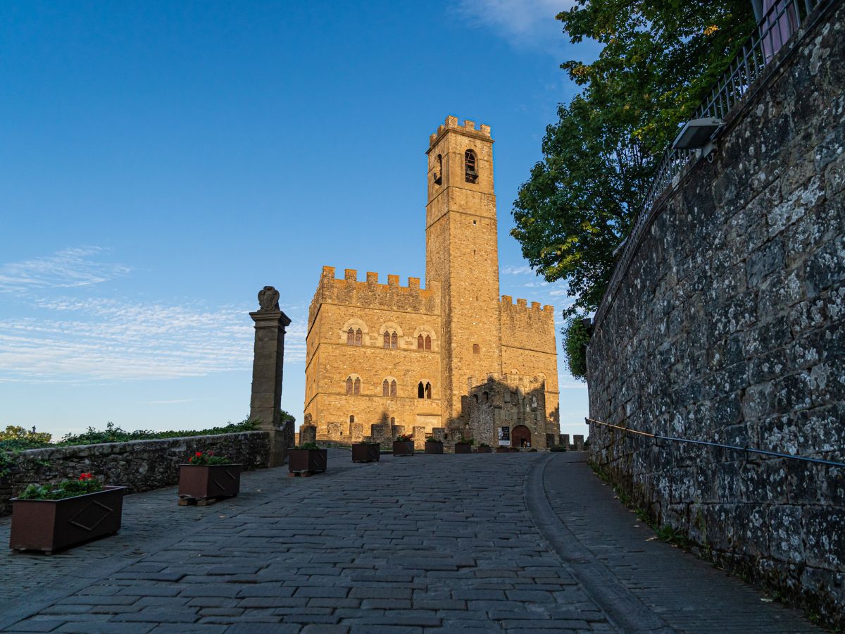 Vista frontale del Castello di Poppi in pietra dorata, con la sua alta torre campanaria che si erge al centro e una strada lastricata in salita in primo piano sotto un cielo azzurro.