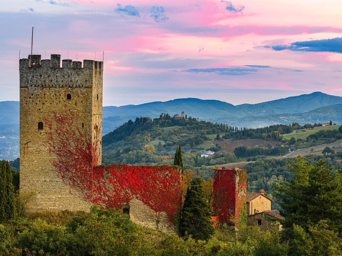 La torre e le mura in pietra del Castello di Porciano parzialmente coperte da edera rossa autunnale, immerse nel paesaggio collinare del Casentino con il cielo sfumato di rosa al tramonto.