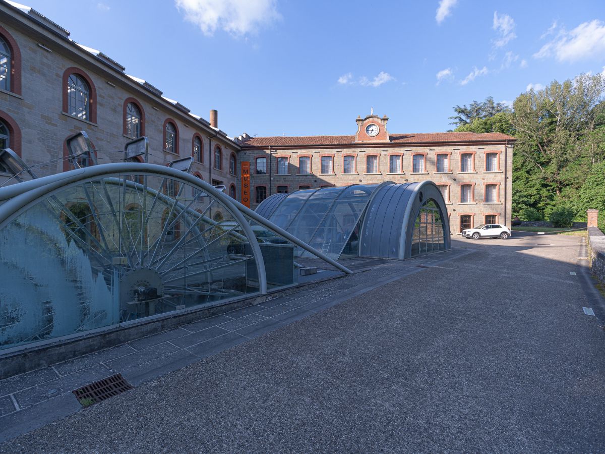 L'esterno del Museo della Lana di Stia, un edificio industriale storico in pietra con ampie strutture moderne a vetrata e una grande ruota ad acqua in metallo.