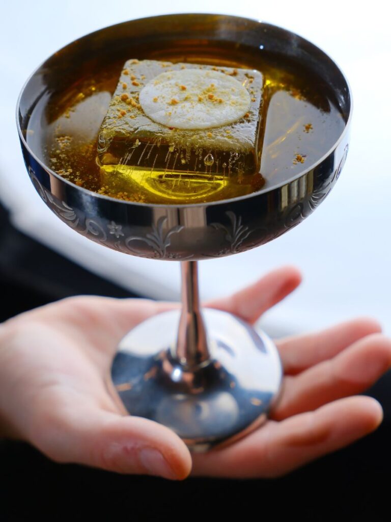 Cocktail artigianale Oro Sacro servito in una coppa di metallo finemente decorata con un cubetto di ghiaccio trasparente.