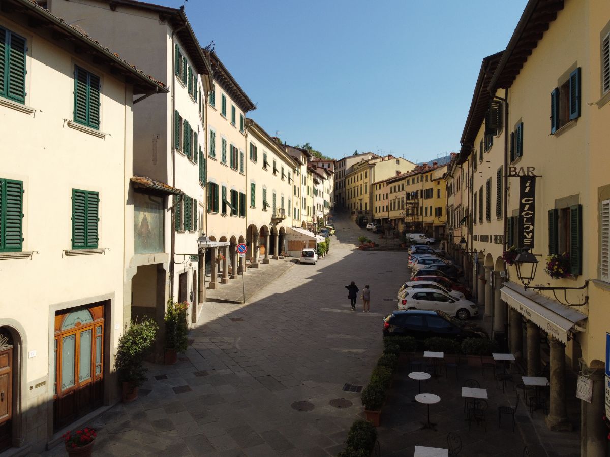 Vista longitudinale di Piazza Tanucci a Stia, una piazza allungata incorniciata da edifici storici con portici, bar all'aperto ed edicole, sotto un cielo sereno.
