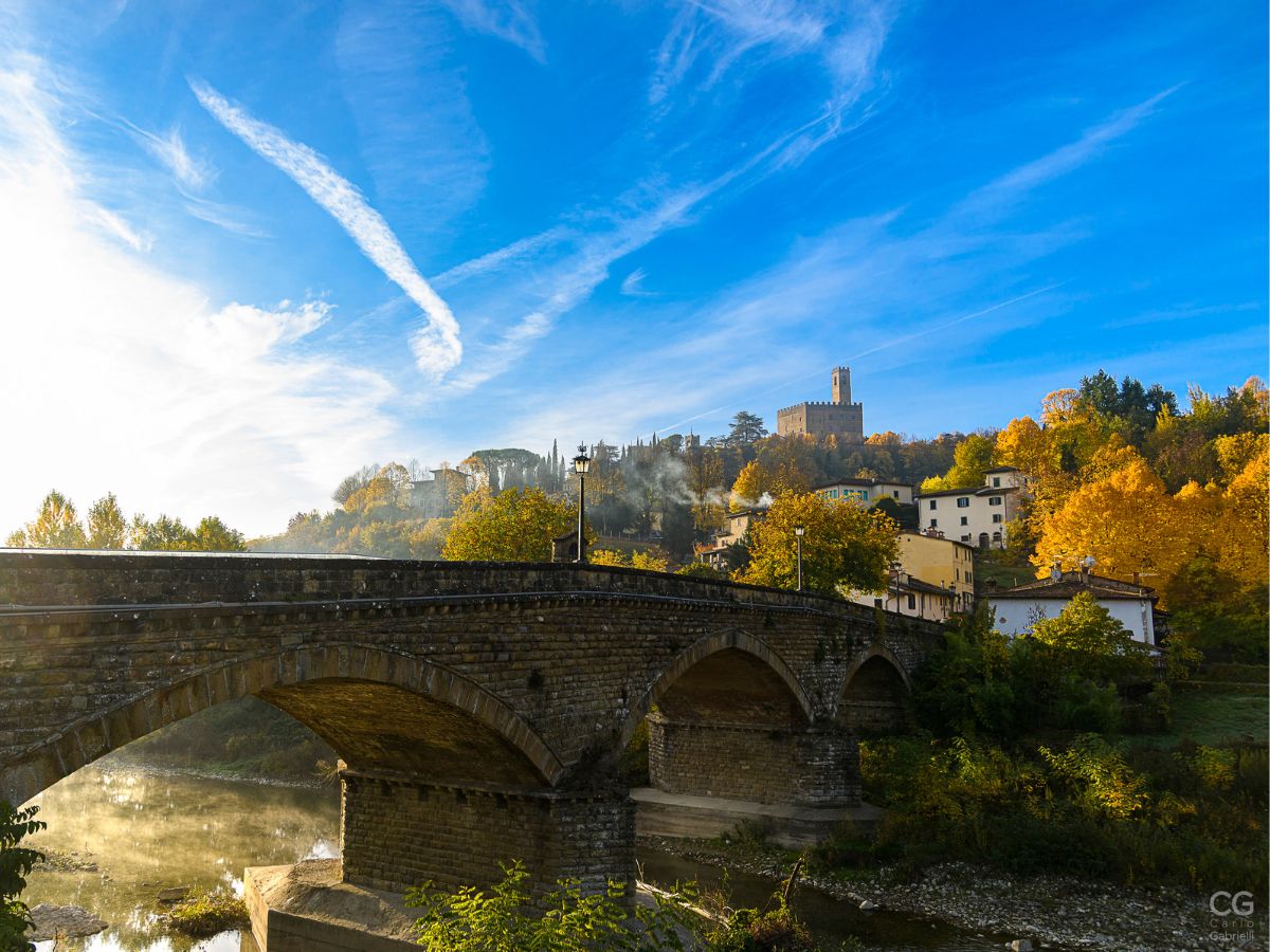 Vista panoramica del Castello di Poppi che svetta sulla collina con il ponte in pietra sul fiume e gli alberi dorati in autunno.