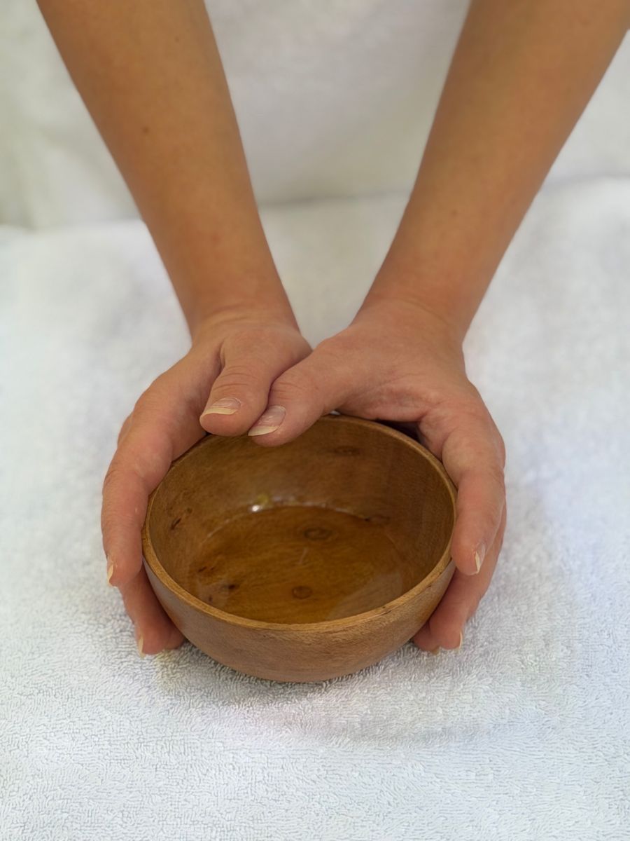 Mani che sorreggono una ciotola di legno con l’olio per il massaggio, su un telo bianco