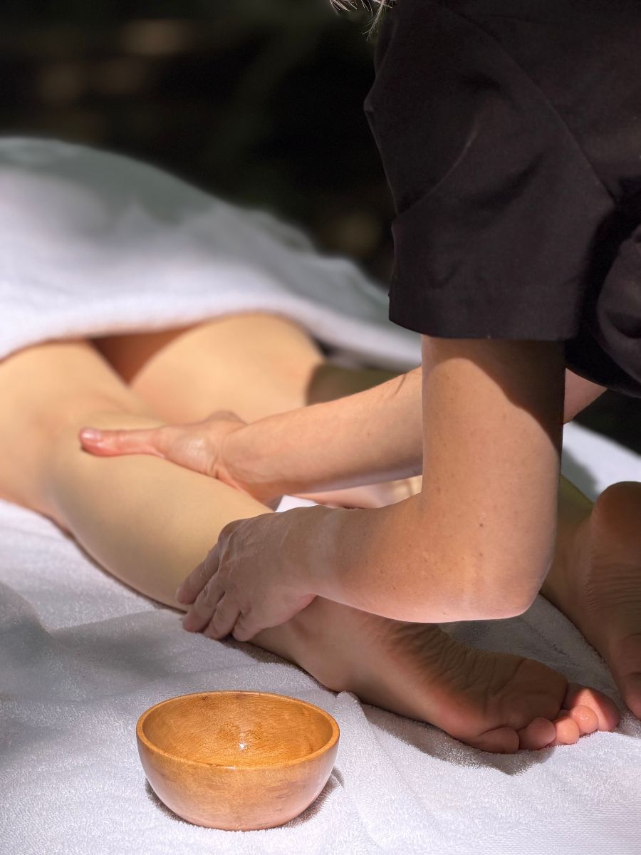 Massaggio alle gambe su lettino con telo bianco e ciotola di legno con l’olio, immersi nel verde